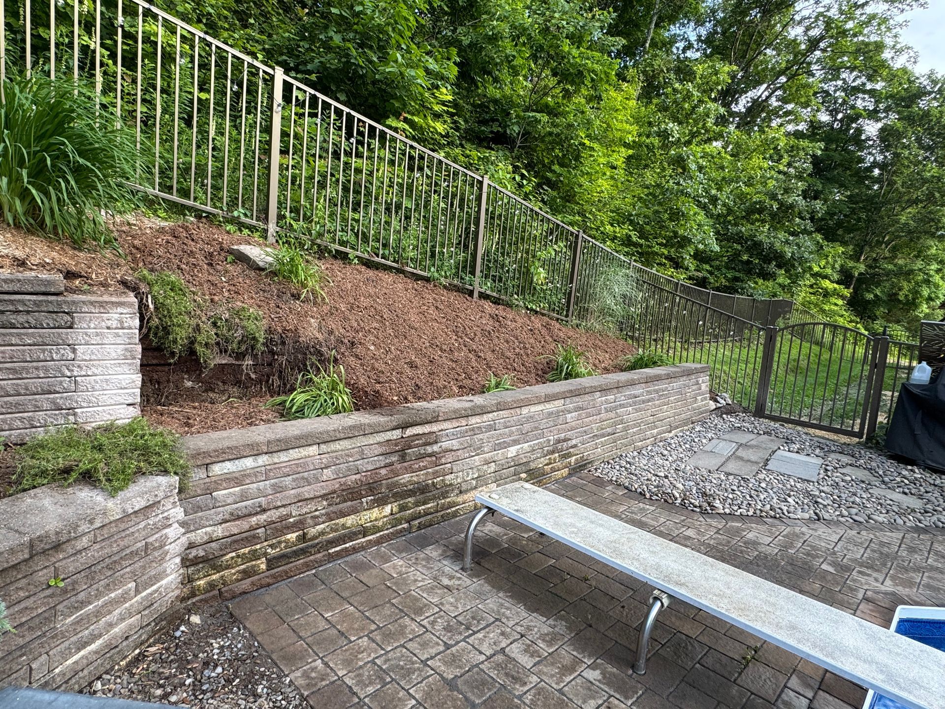 A sloped backyard with a retaining wall, fence, and a paved patio. Mulch covers the slope, with trees in the background.
