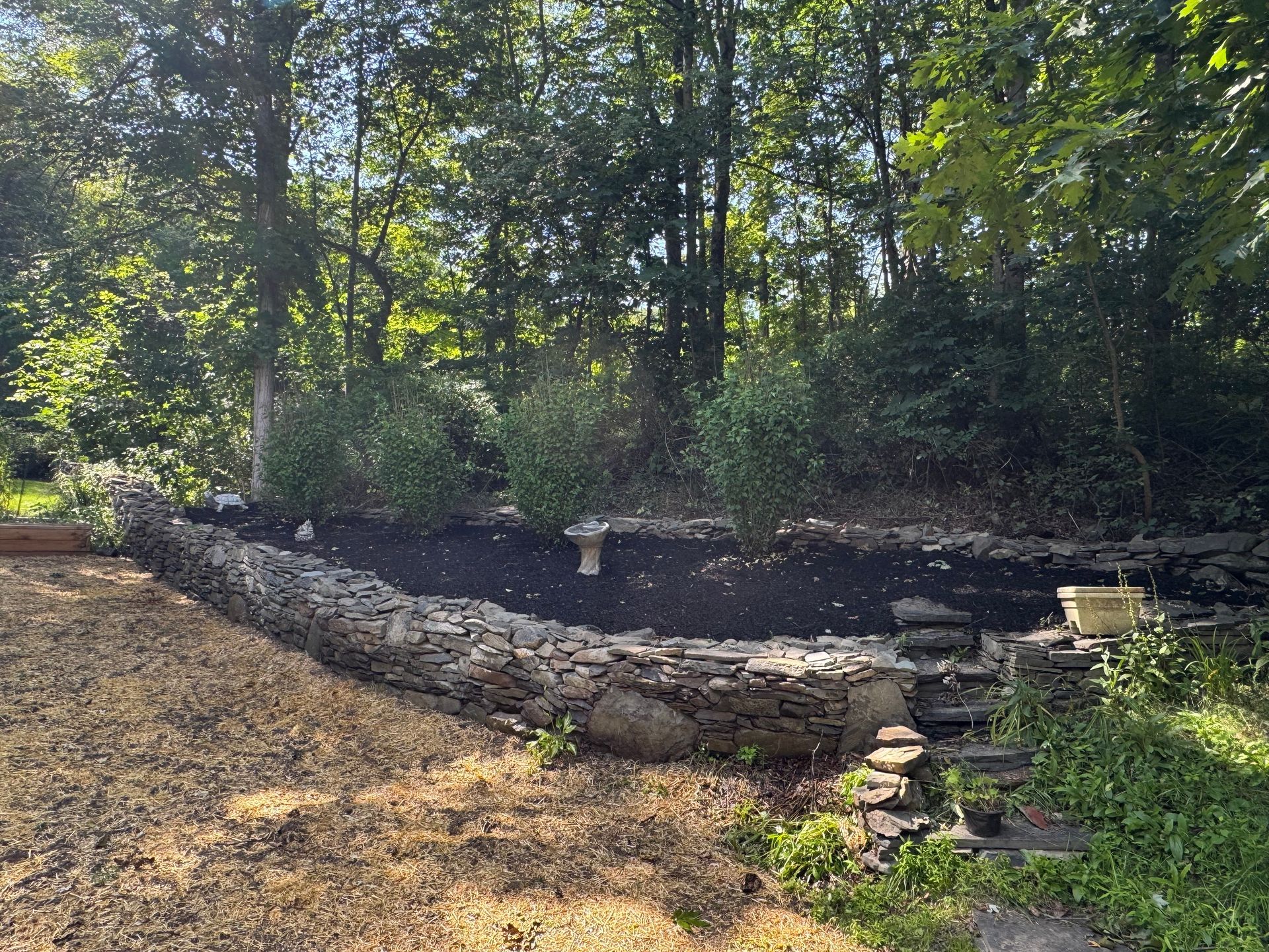 Stone-walled garden bed with mulch and shrubbery. A small set of stone steps leads up to the bed. Background is a wooded area.