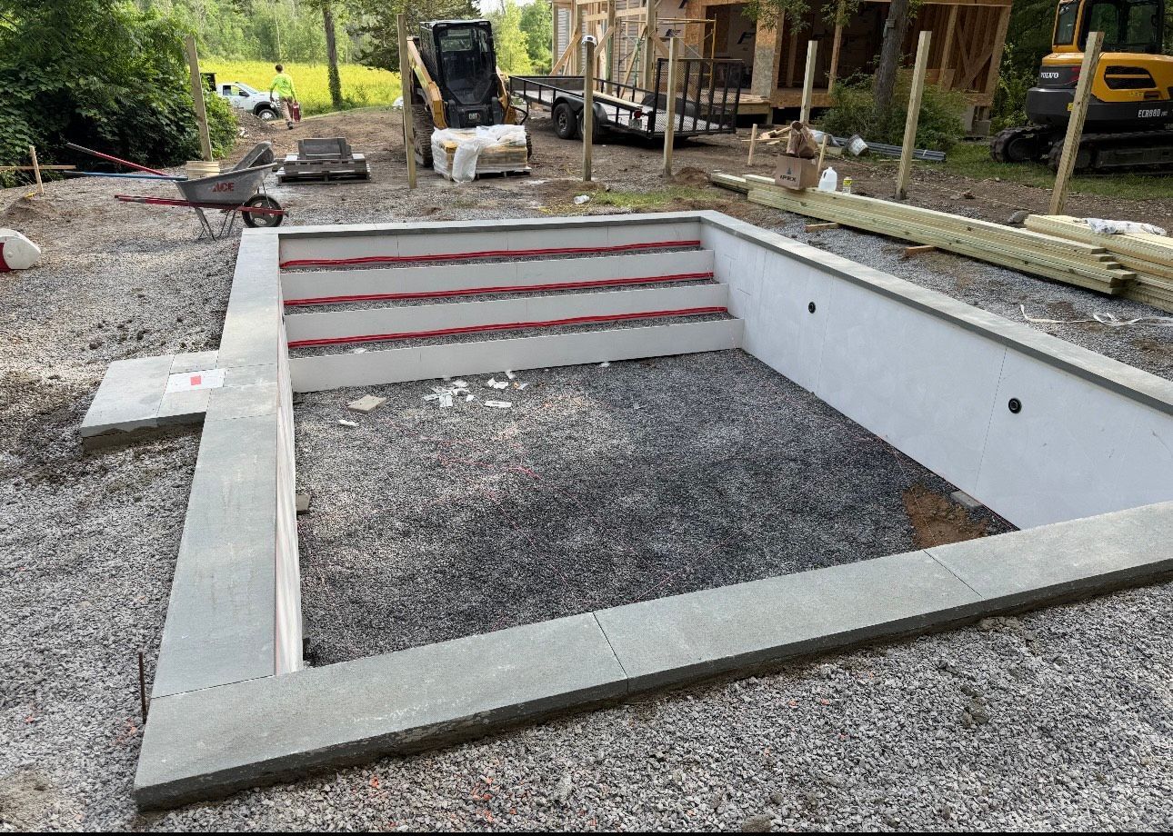 A rectangular pool under construction, with gray concrete walls, a step entry, and gravel inside. Construction equipment and materials are visible in the background.