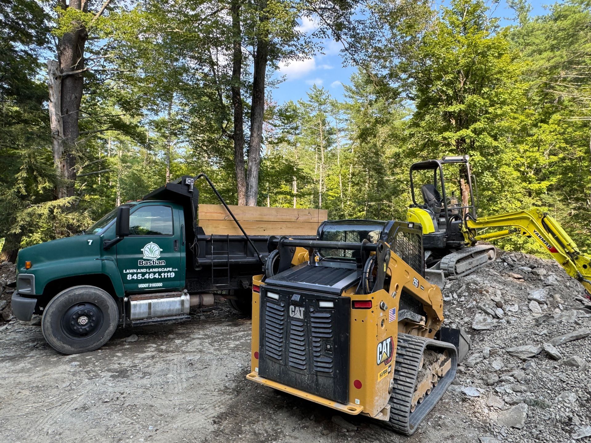 A green dump truck, yellow skid steer, and excavator near a gravel pile in a wooded area.