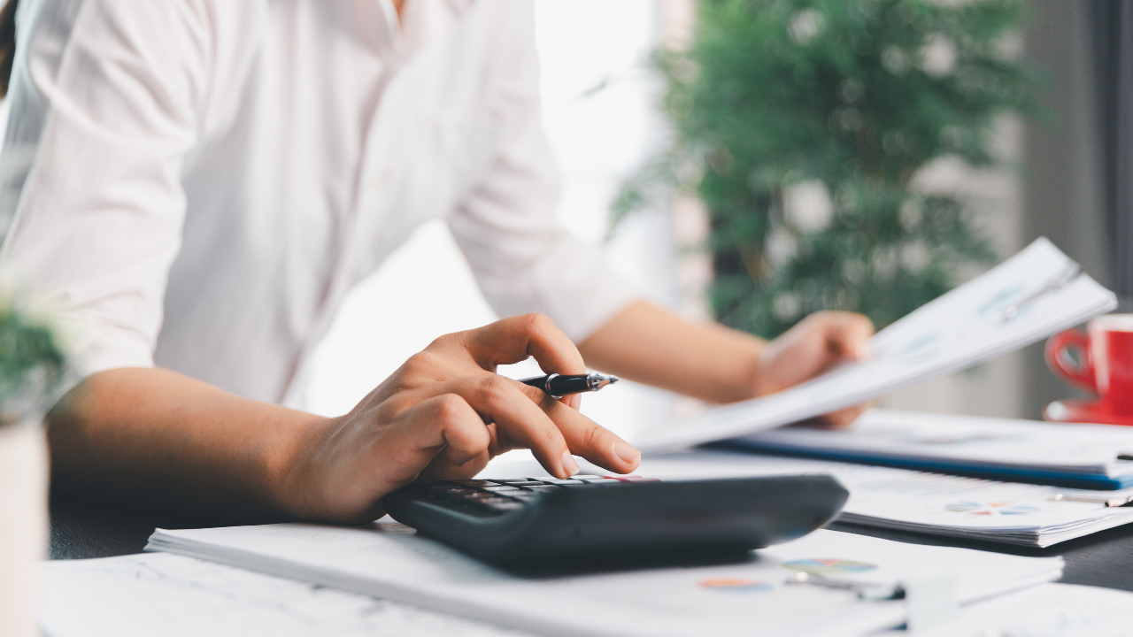A person in a white shirt sits at a desk using a calculator while holding documents.