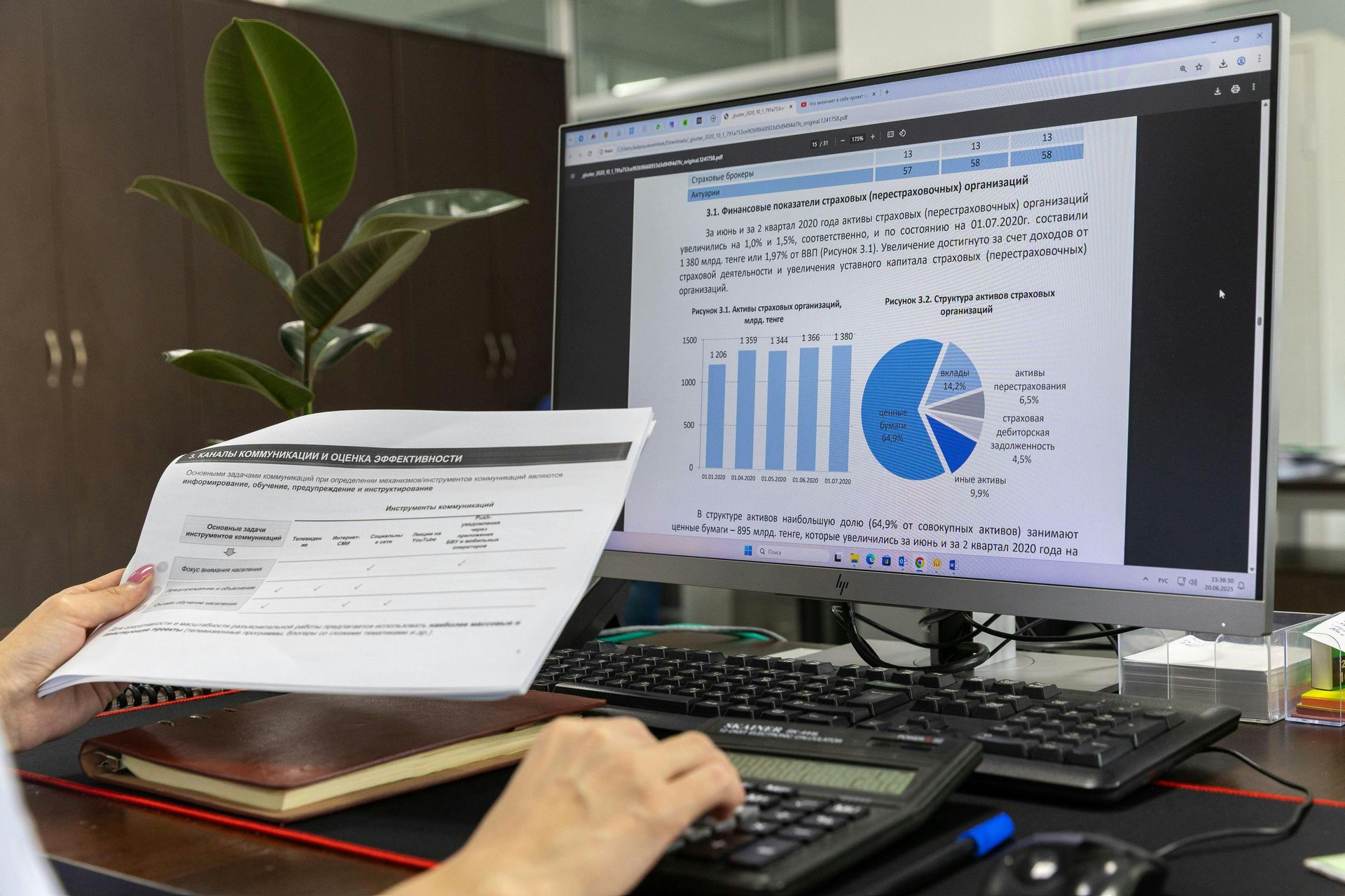 A person calculates figures using a document and a computer screen displaying a bar chart and a pie chart in an office.