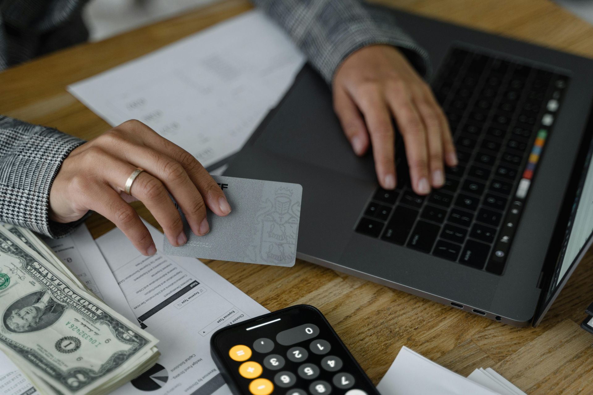 A person uses a laptop and credit card while working with cash and documents on a desk with a calculator nearby.