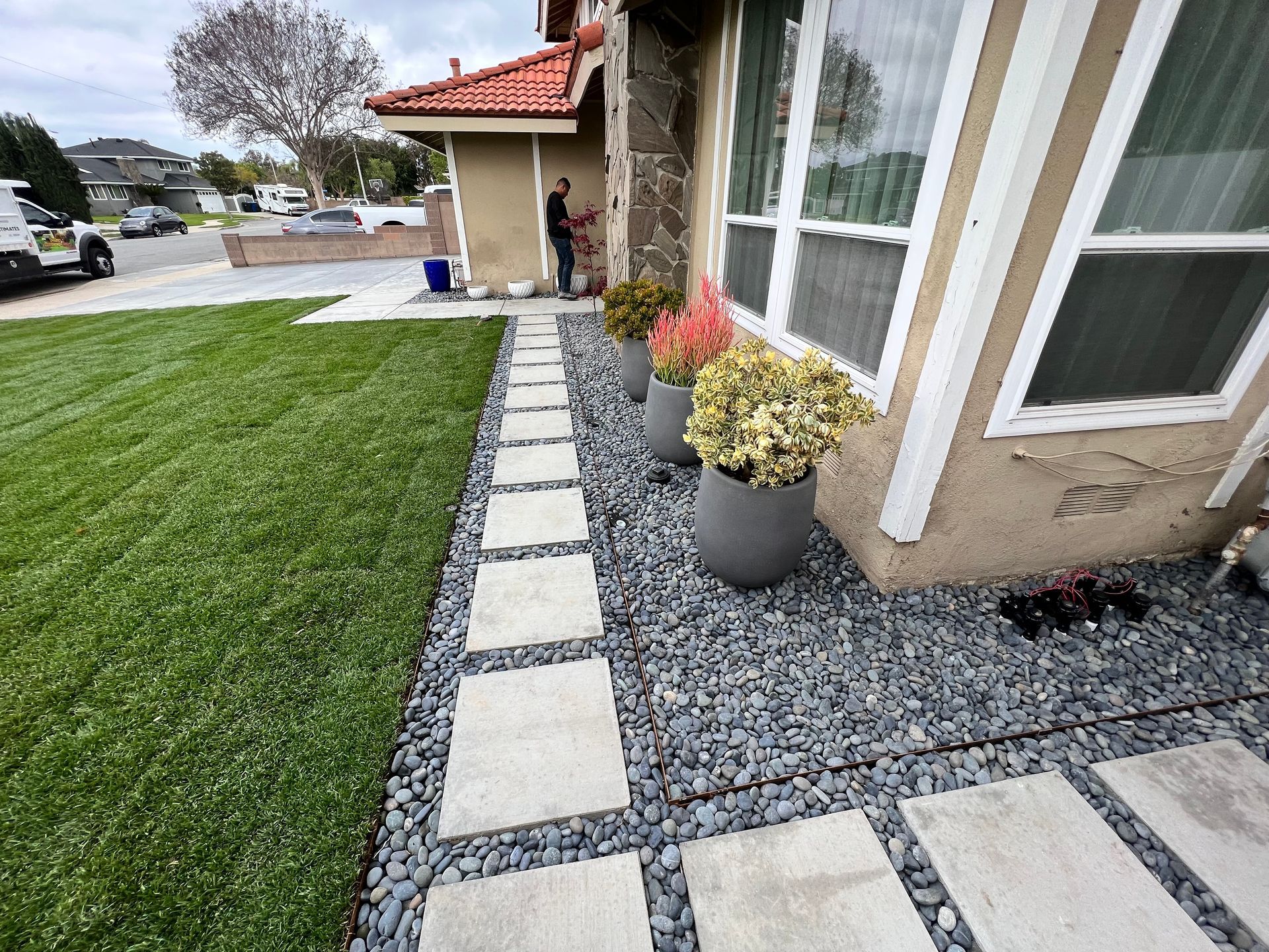 Front yard walkway with square stepping stones, green lawn, gravel bed, and potted flowers by a house entrance