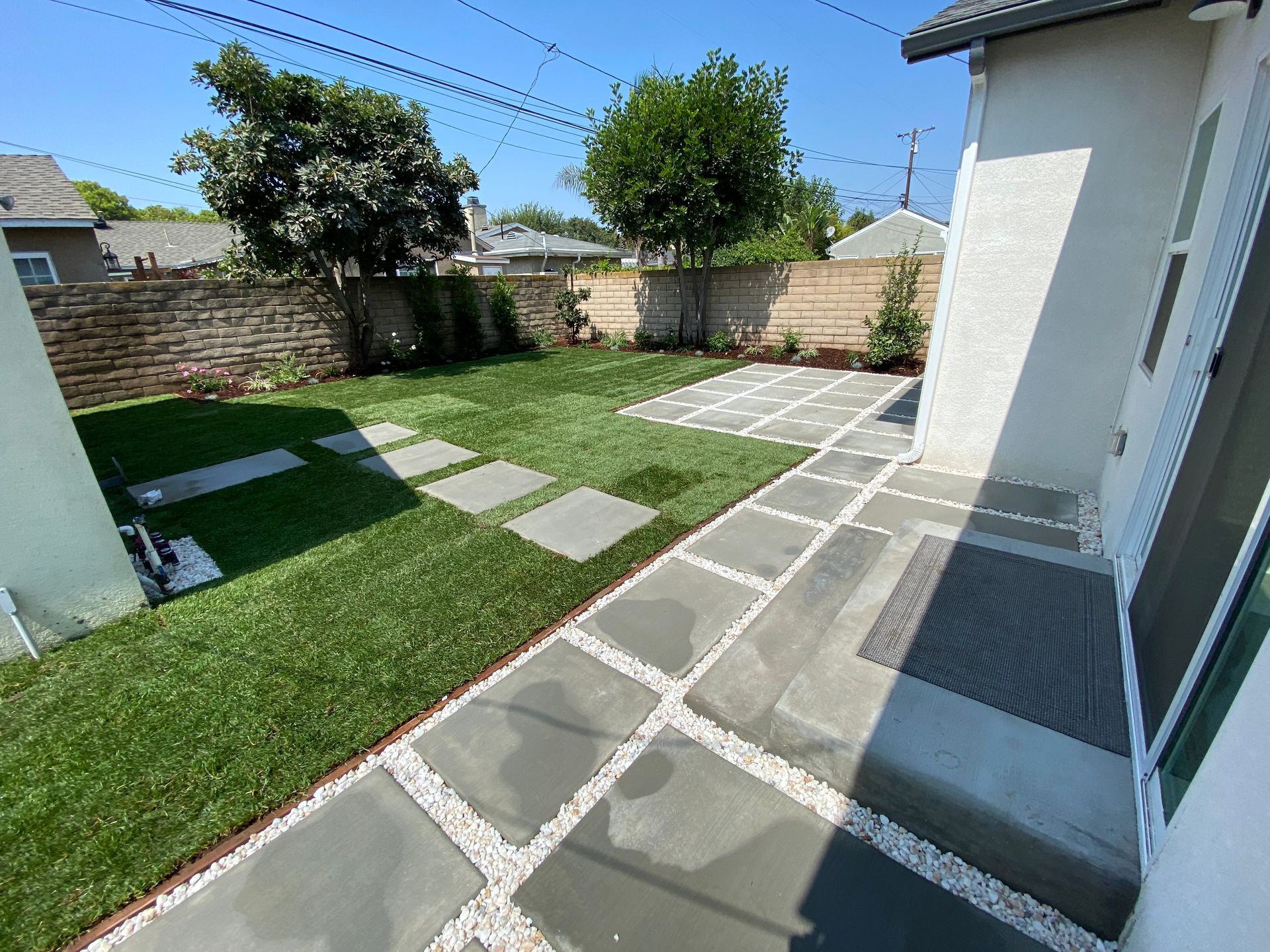Backyard patio with gray stone pavers, green lawn, and a white house wall under a clear sky