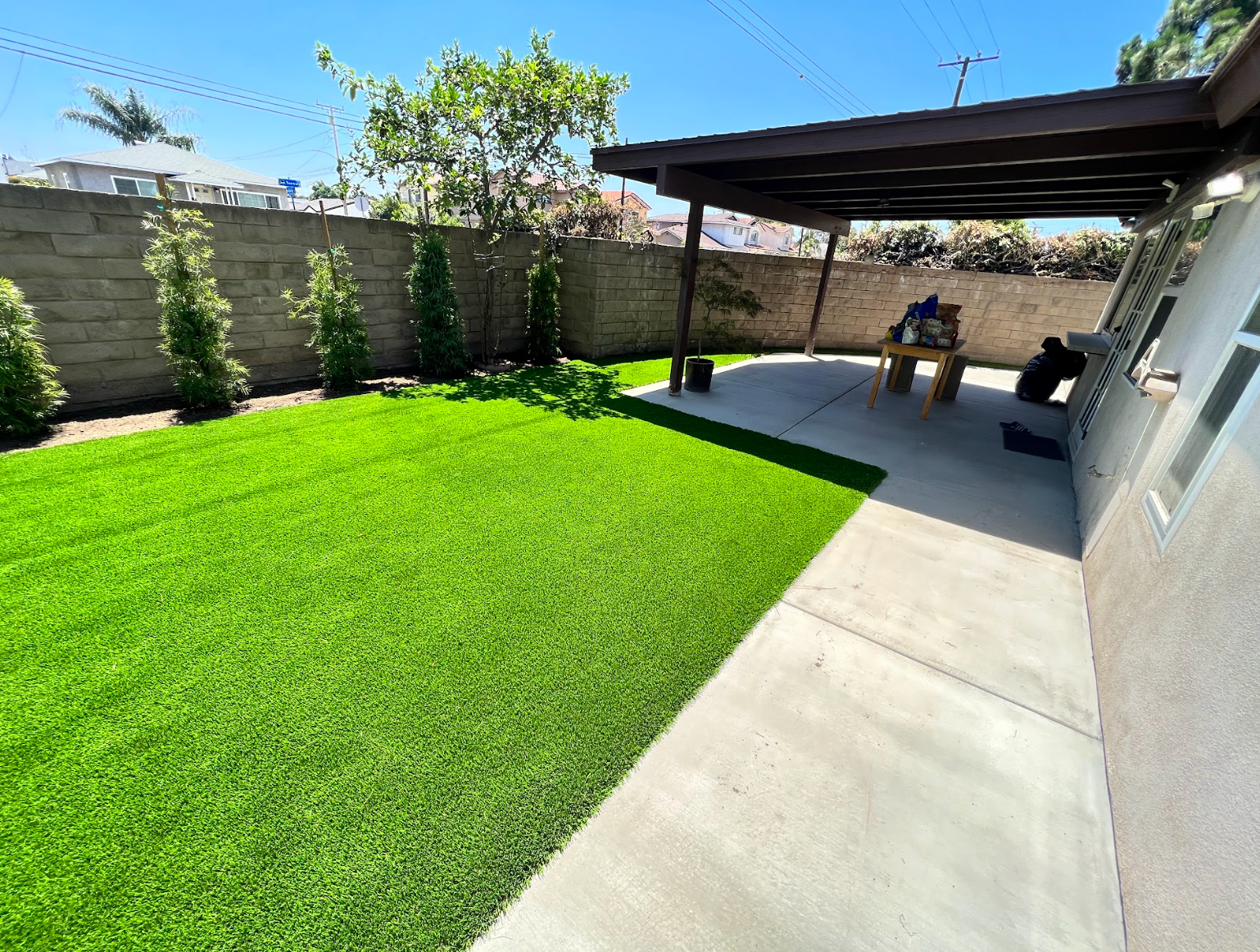 Backyard patio with bright green lawn, concrete walkway, and covered seating area by a wall
