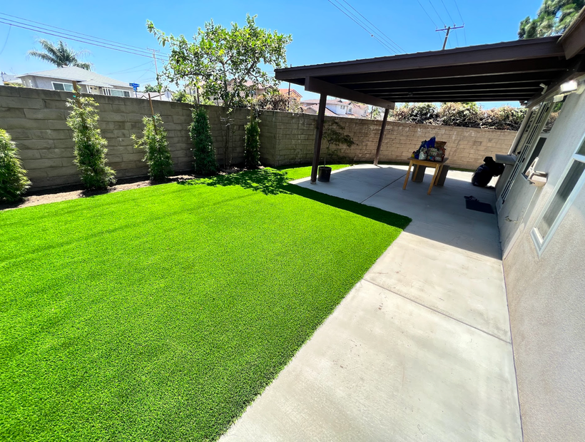 Backyard patio with bright green lawn, concrete walkway, and covered seating area by a wall