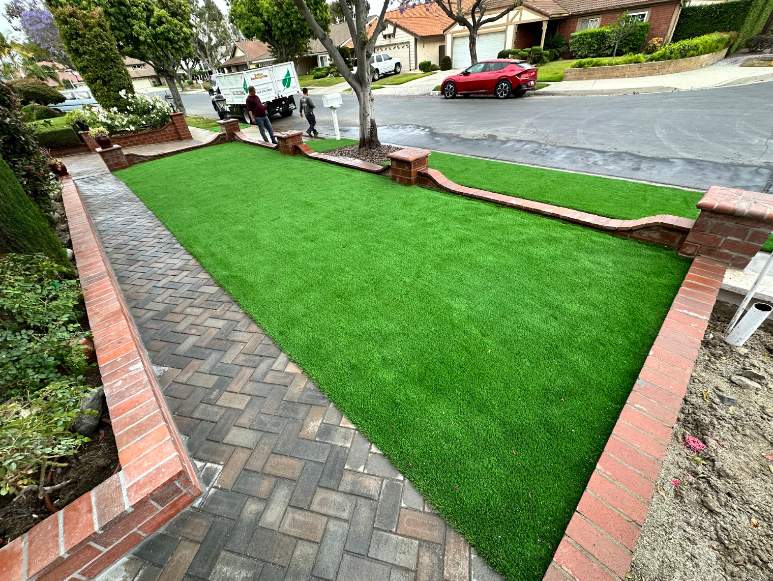 Brick walkway beside a bright green lawn leading to a residential street with a red car.