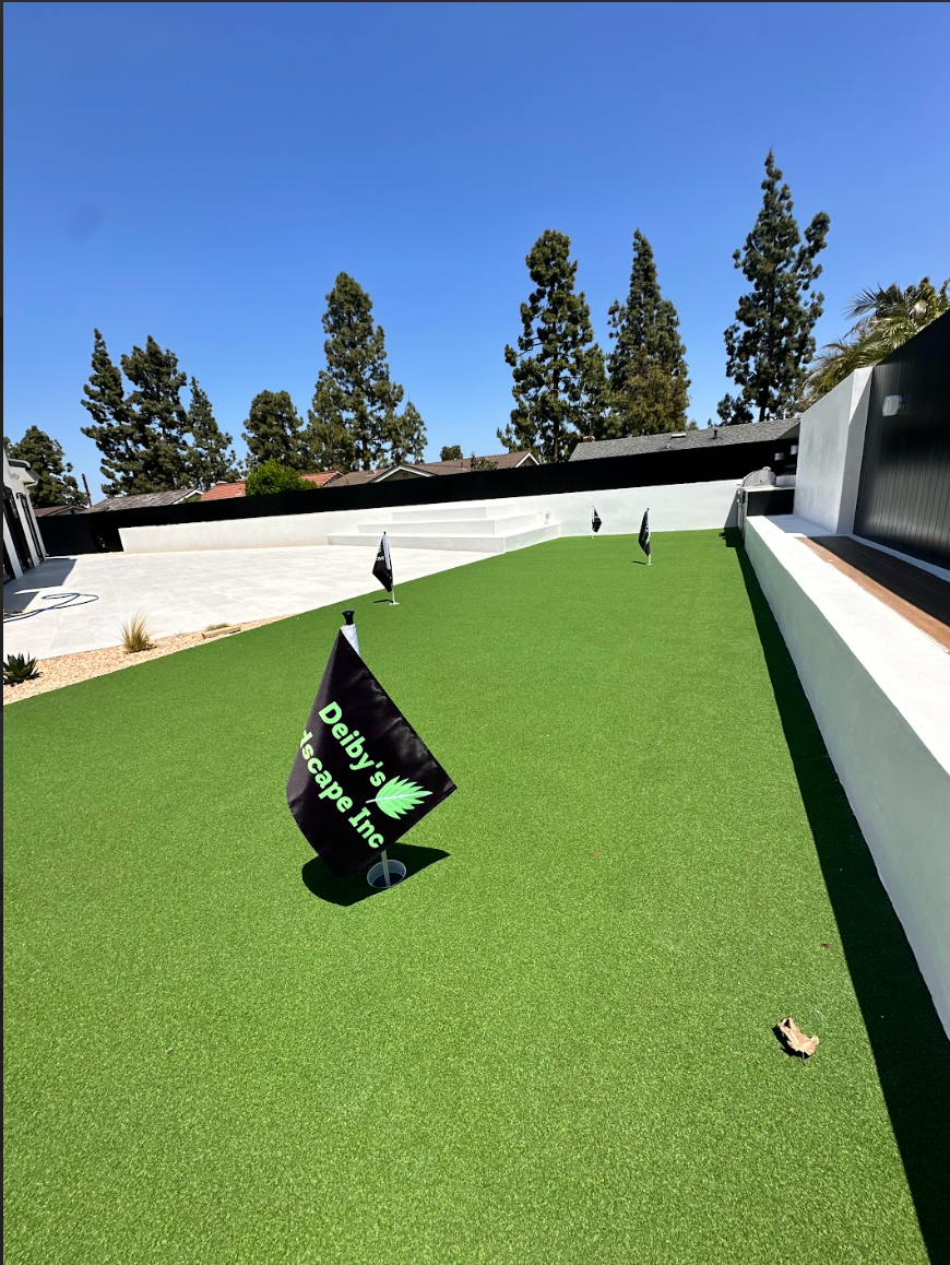Small rooftop putting green with a black flag, white walls, and trees under a clear blue sky