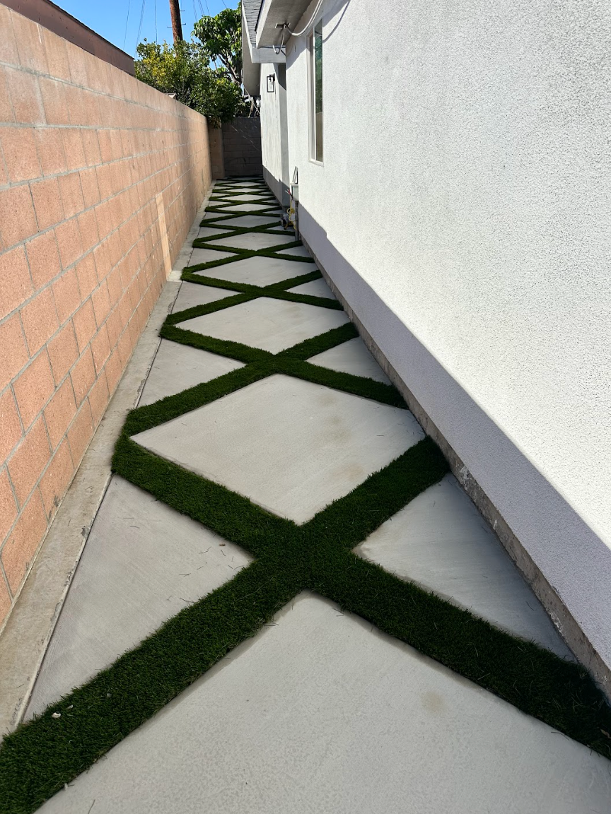 Narrow side yard walkway with white pavers and green grass X-patterns between a stucco wall and a brick wall