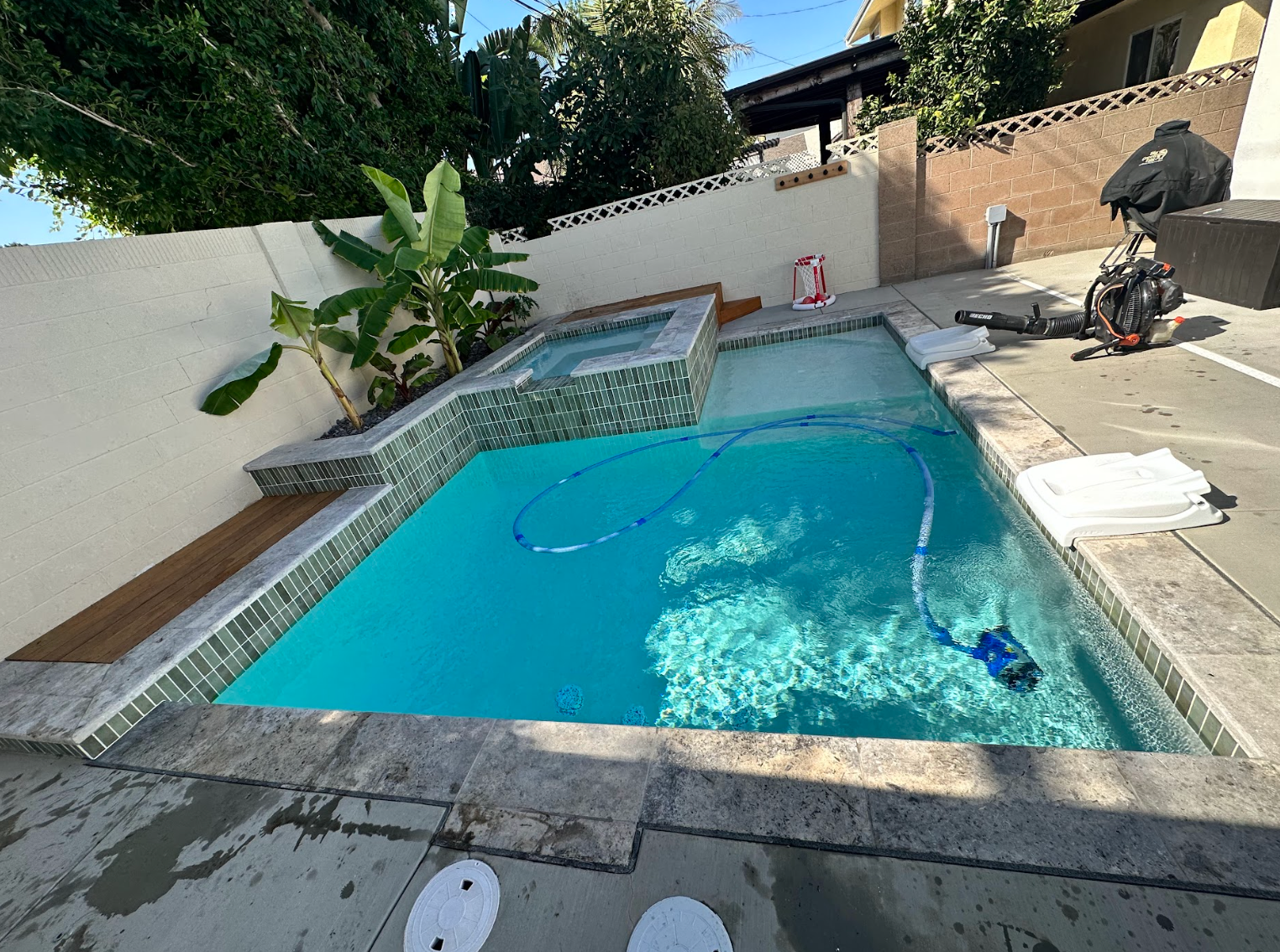 Backyard swimming pool with turquoise water, tiled deck, lounge chair, and potted plants in the sun