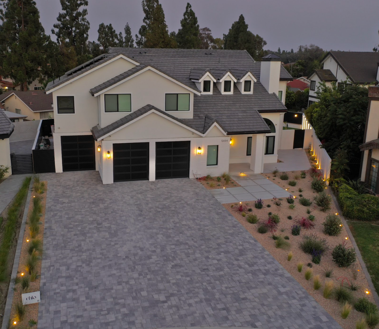 Modern two-story suburban house with lit garages, large driveway, and landscaped front yard at dusk