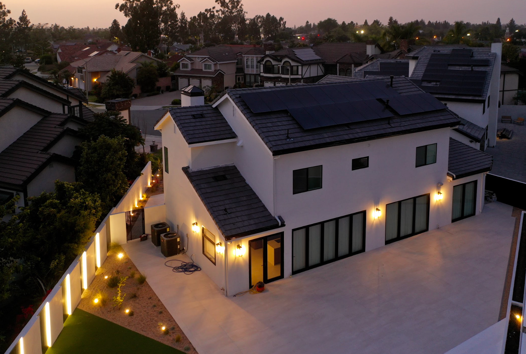 Modern white house with solar panels and warm lights at dusk, viewed from above in a residential neighborhood
