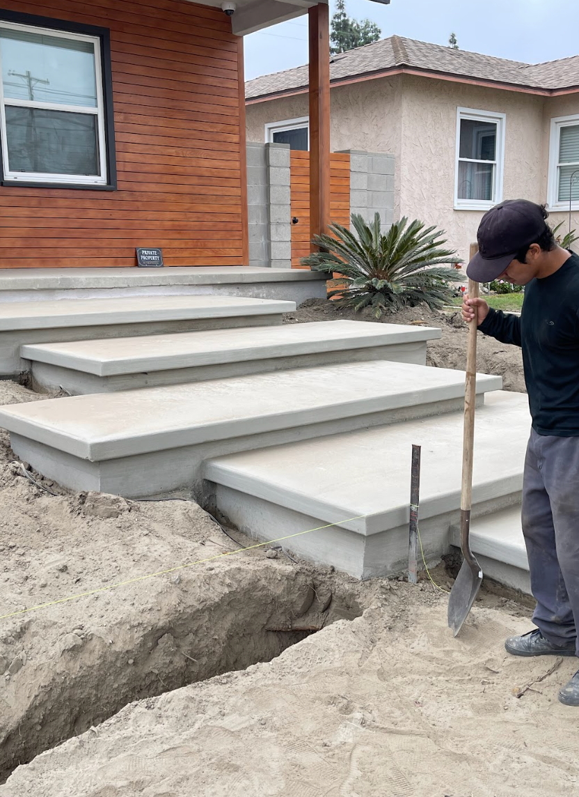 Worker measuring a concrete front step and walkway beside a brick house and dirt excavation.