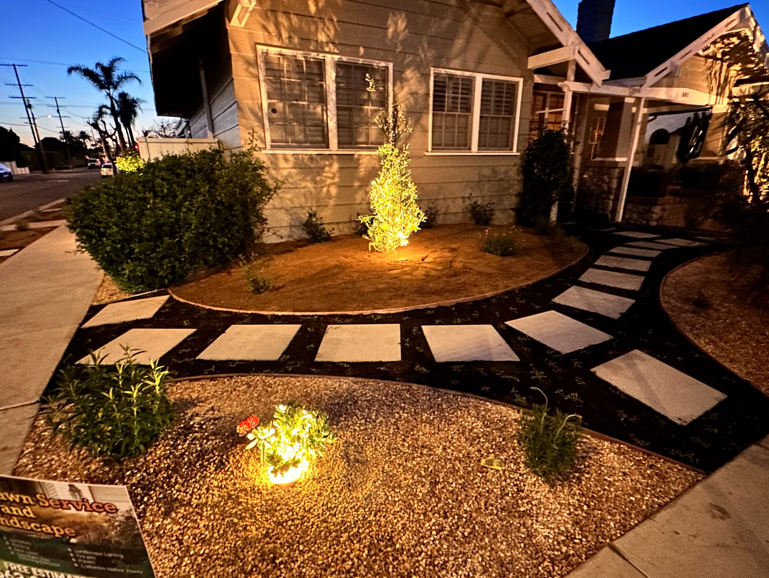 Front yard landscape with stepping stones, lit plants, and a house at dusk