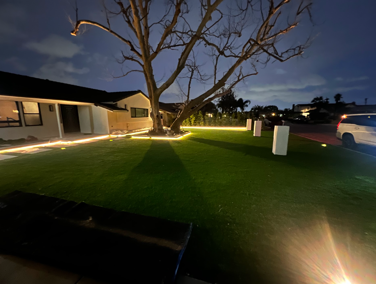 Nighttime suburban front yard with a large tree, lit lawn, and a parked car at right