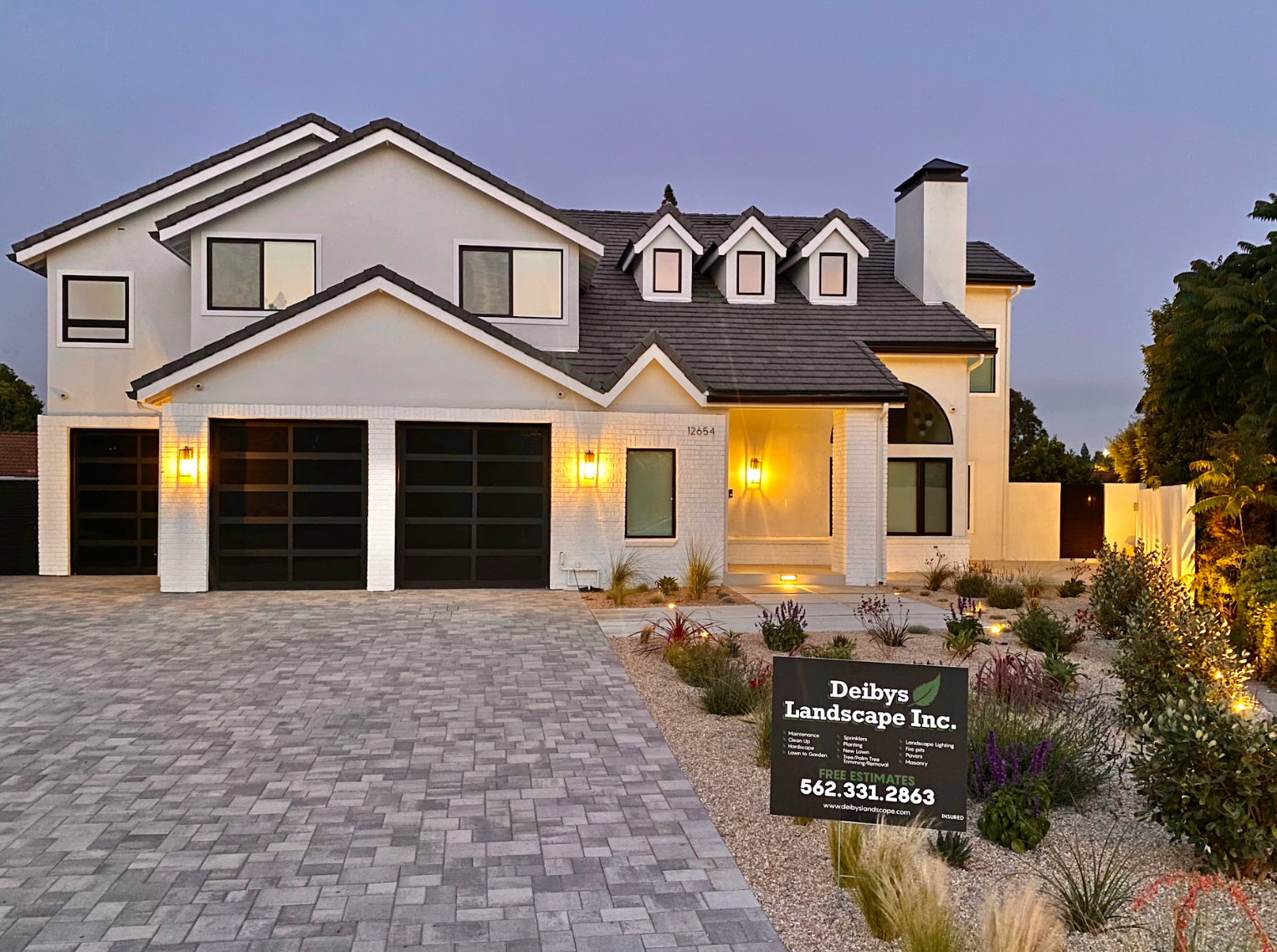 Luxury two-story house with lit windows, three-car garage, and stone driveway at dusk