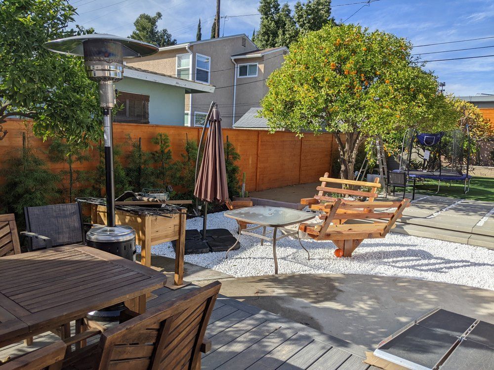 Backyard patio with wooden tables, benches, umbrella, heater, and trees beside a wooden fence