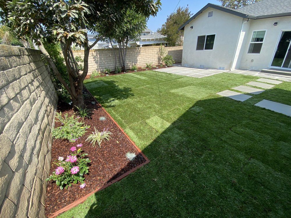 Small backyard with green lawn, stone wall, flower bed, and a white house patio.