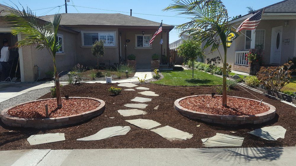 Front walkway with stone pavers between two circular mulch beds and small palm trees in front of homes