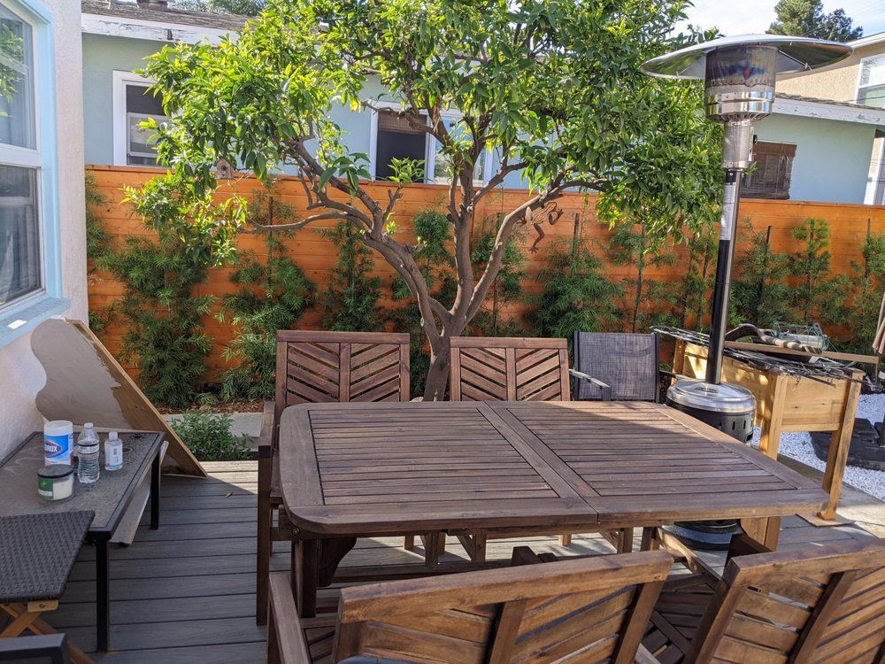 Outdoor patio with wooden table and chairs, potted trees, and a brick privacy wall in sunlight