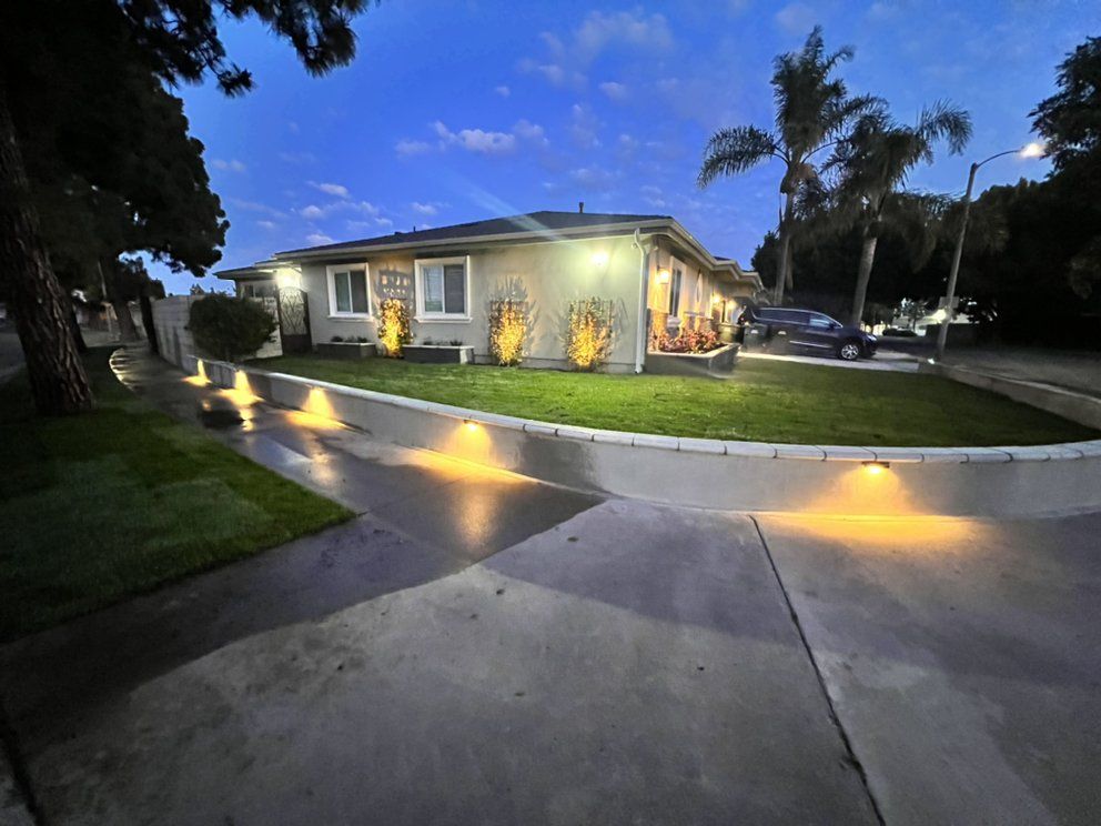 Residential street corner at dusk with lit single-story house, glowing path lights, and palm trees.