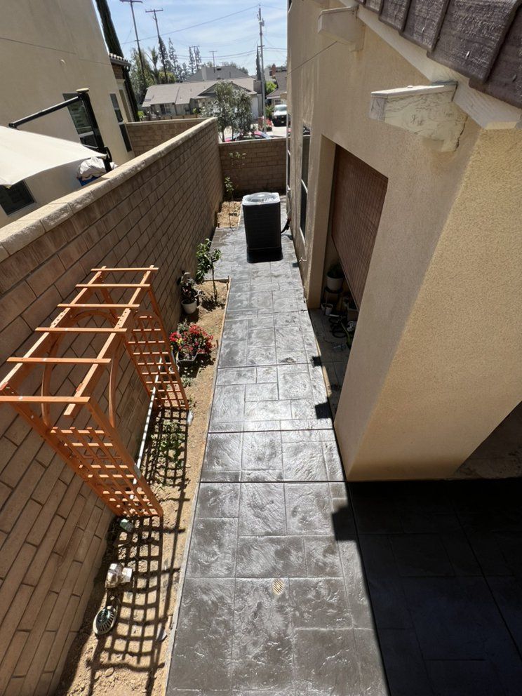 Narrow outdoor walkway between buildings with gray stone pavers, tan walls, and a wooden trellis on the left.