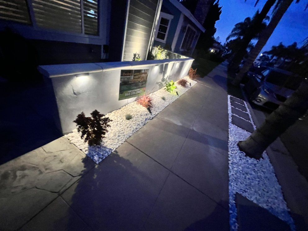 Dimly lit house exterior with a concrete walkway, white stone border, and small plants along the wall at dusk