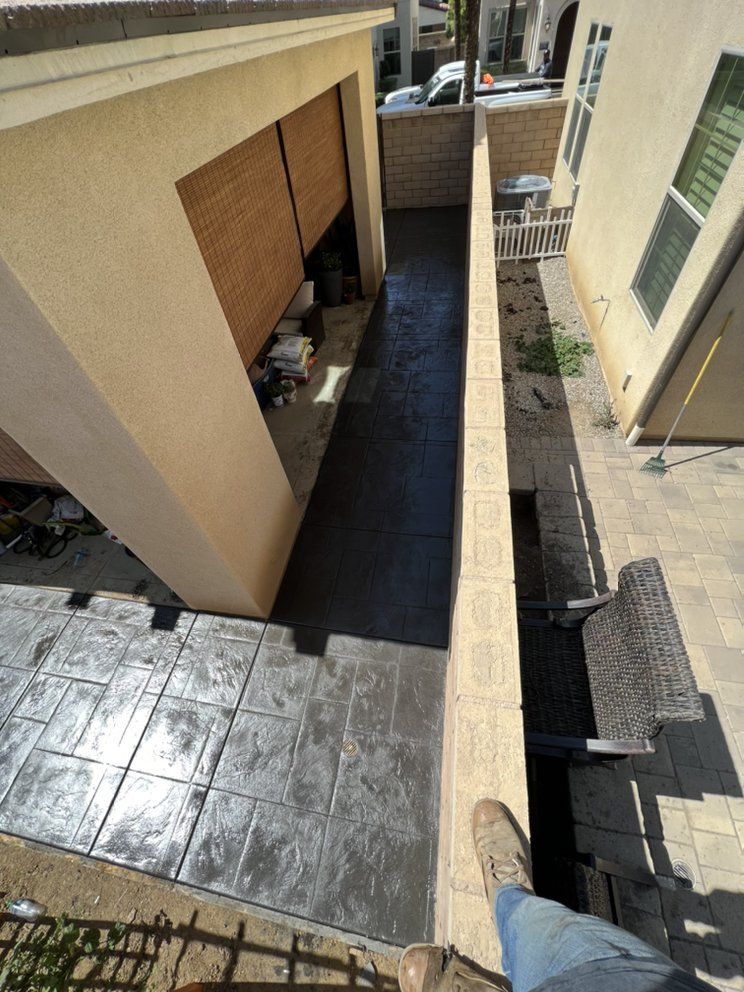 Narrow outdoor walkway beside beige buildings, with tiled ground and a person’s feet at the edge.
