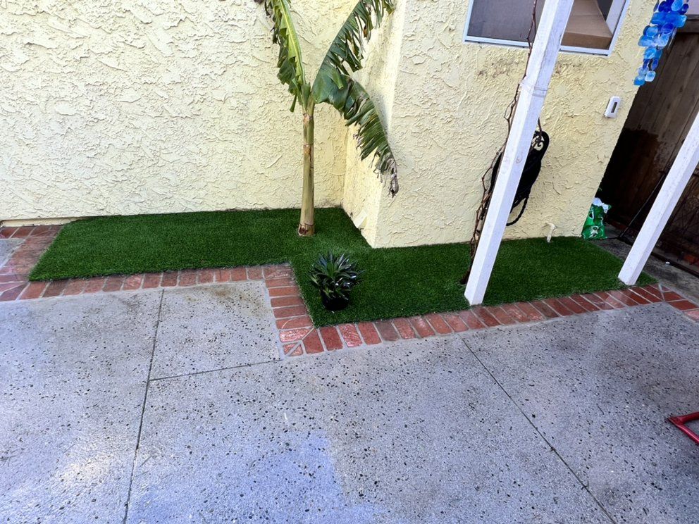 Concrete patio beside a yellow house, with a brick border, small palm tree, and white support posts.