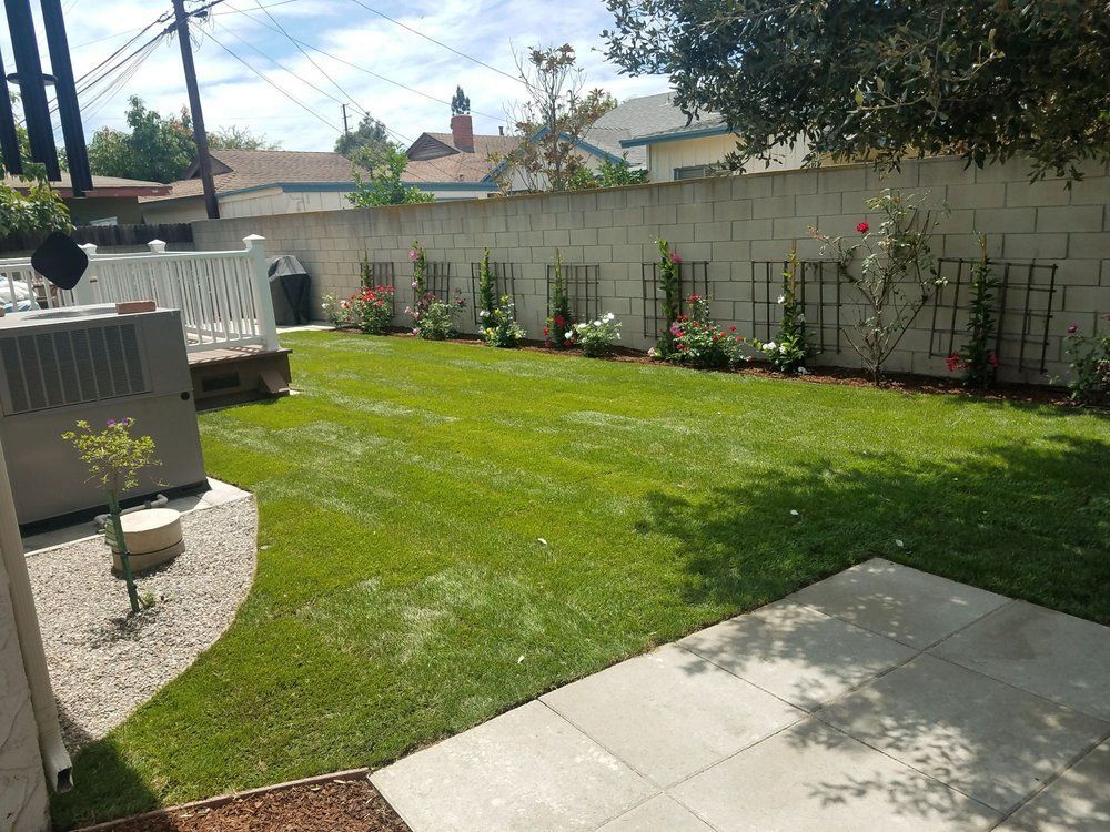 Backyard lawn with patio, planter beds, white fence, and a paved seating area in sunlight