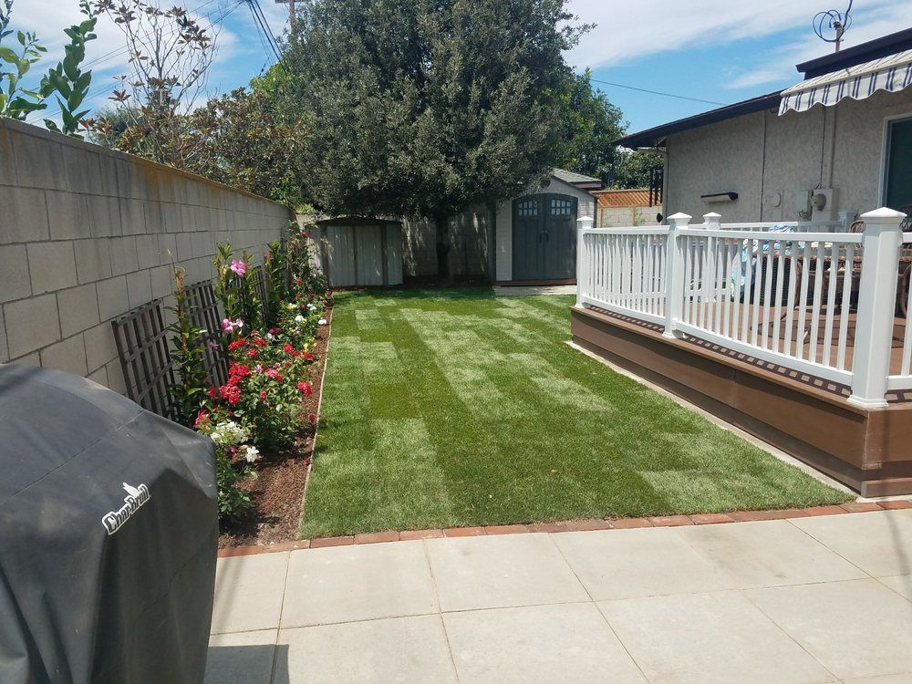 Backyard patio with green lawn, flower bed, grill, and white deck railing under a blue sky