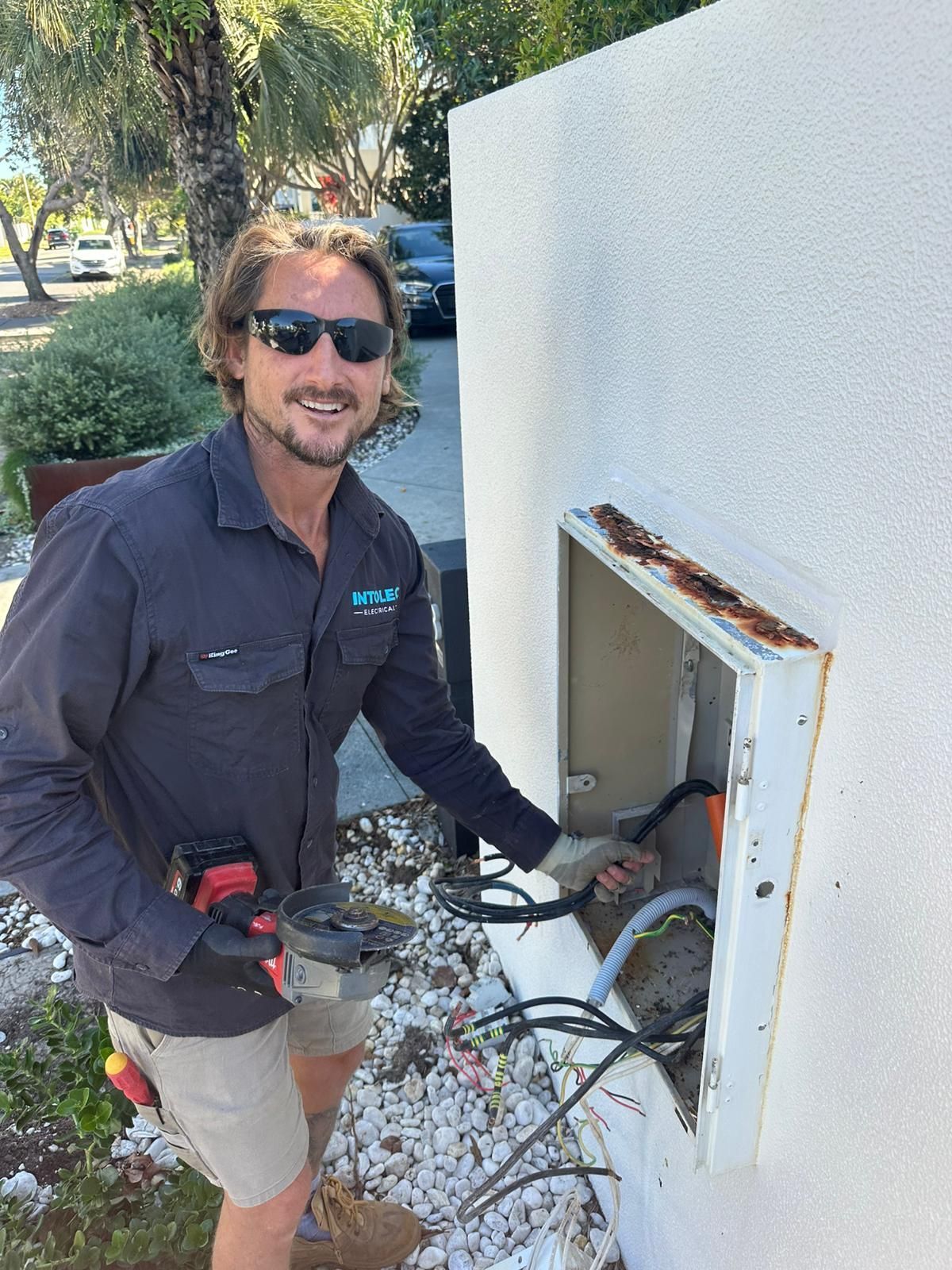 Electrician working on outdoor wiring, holding tool, smiling. White building, sunny day.