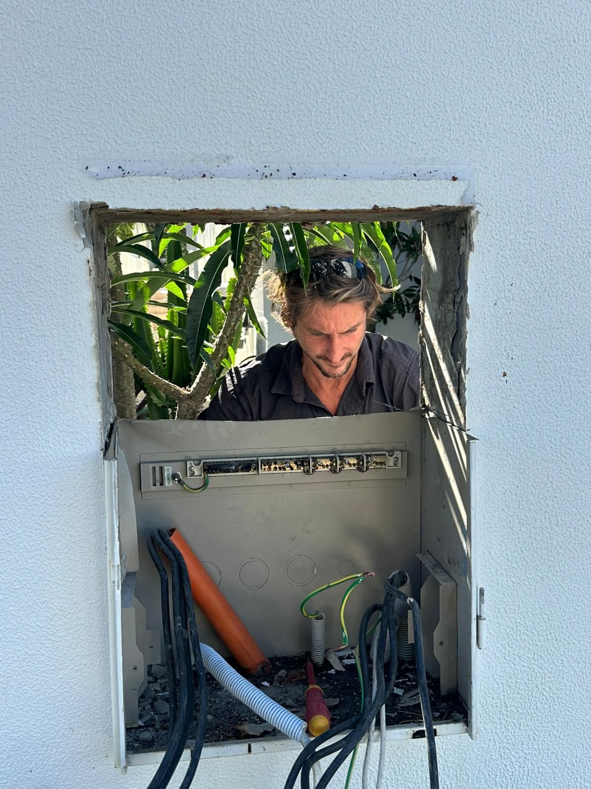Man working on electrical box in a wall, outdoors. Box open, with wires and tools visible.
