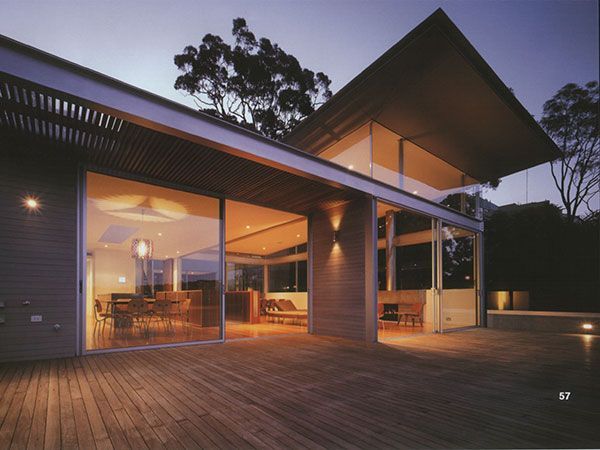 Modern house with large windows, wooden deck, and overhanging roof, illuminated at dusk.