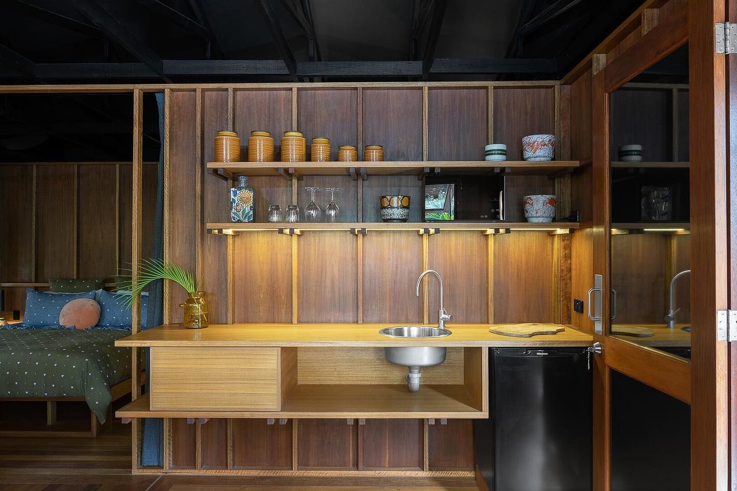Wood-paneled kitchenette with sink, shelves of ceramics, and open doorway to a bedroom with green bedding. — Intolec Electrical In Tweed Heads, NSW