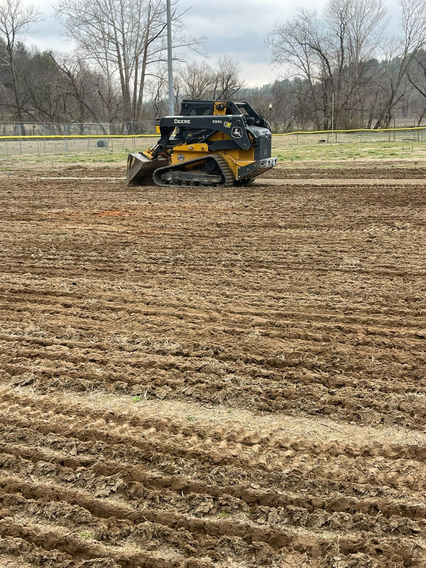 Yellow skid steer in a plowed field with bare trees in the background