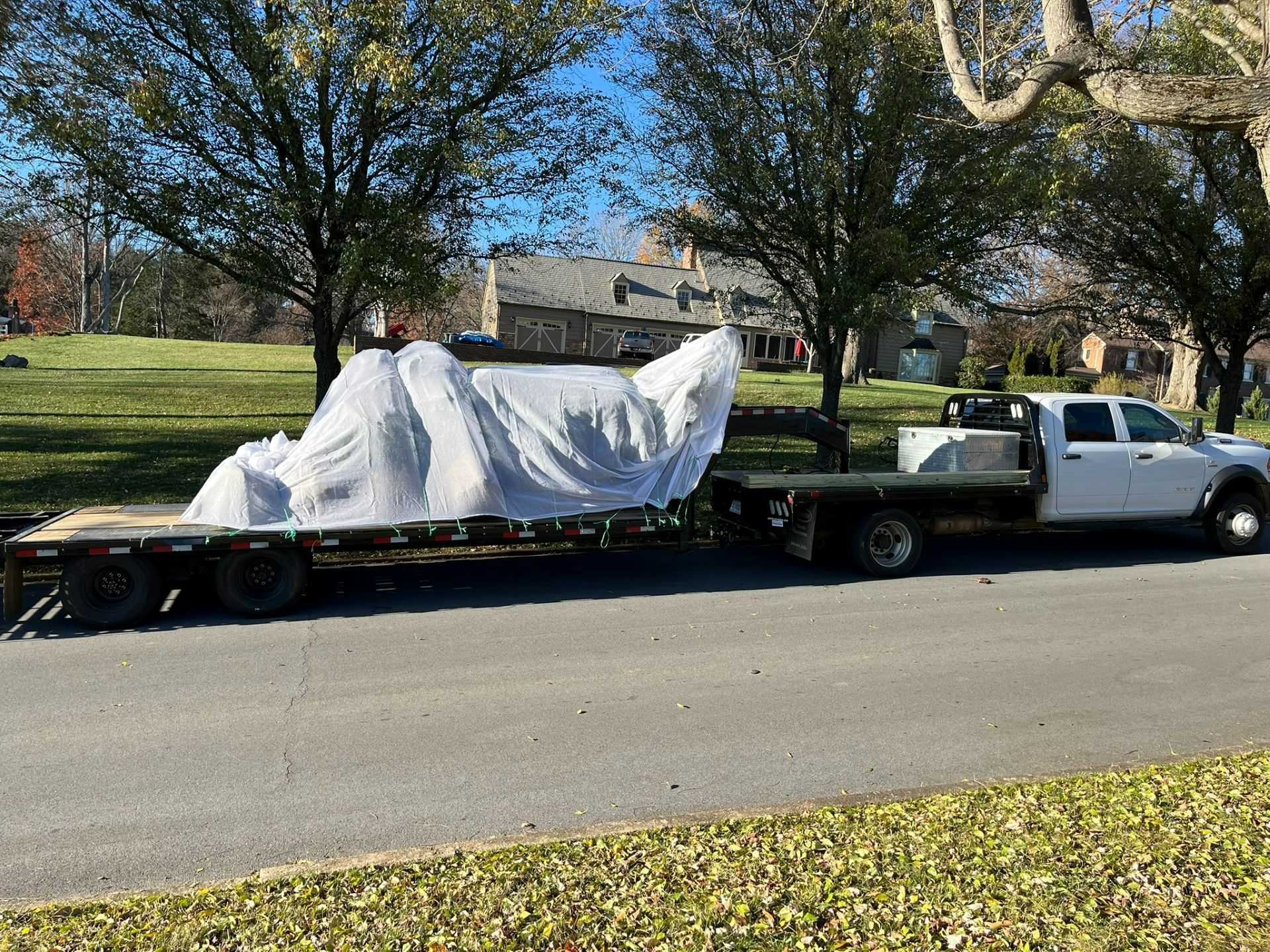 White-covered furniture on a flatbed trailer being towed by a white pickup truck on a suburban street.