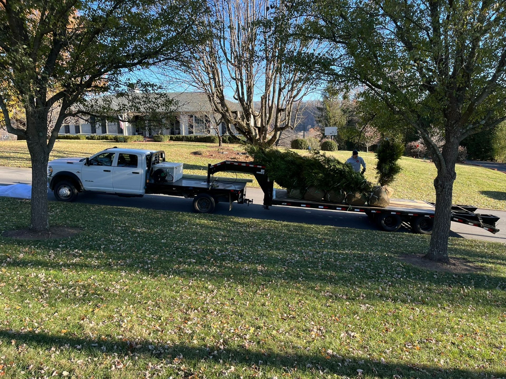 White pickup truck hauling equipment on a flatbed trailer in a grassy park with trees and houses nearby