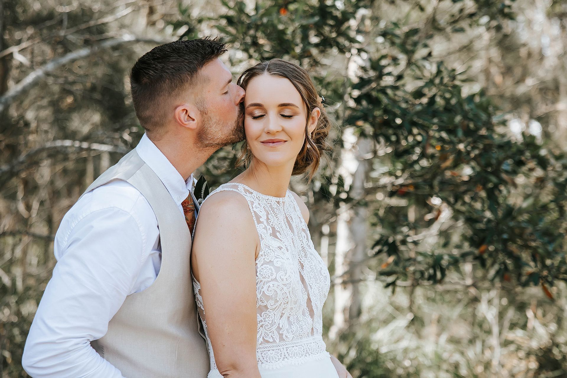 Groom kissing bride's forehead; both are smiling. They stand near trees in Northern NSW