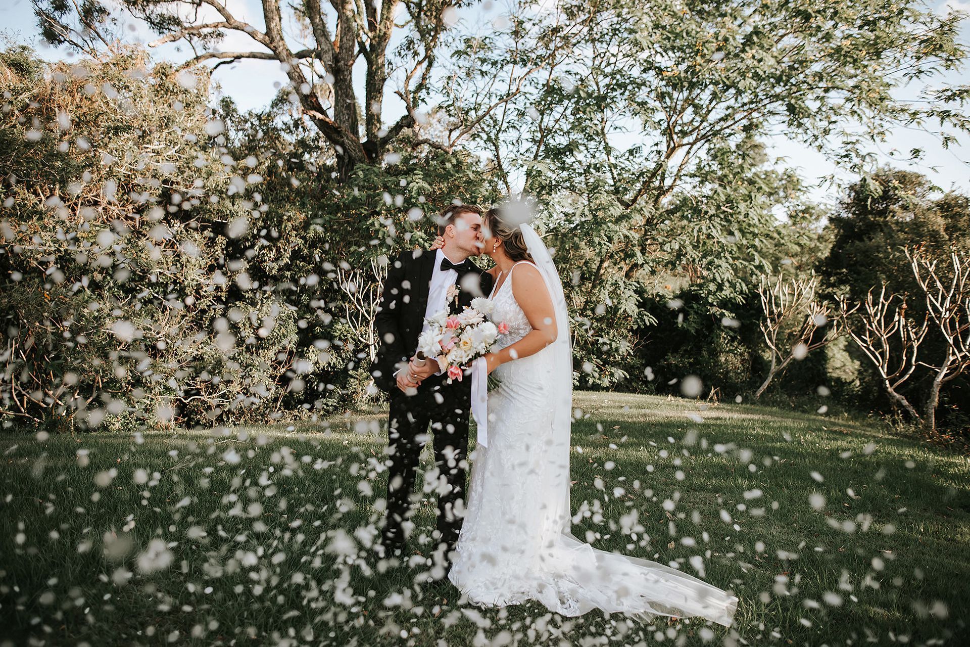 Newlyweds kiss outdoors in Northern NSW, surrounded by falling white confetti.