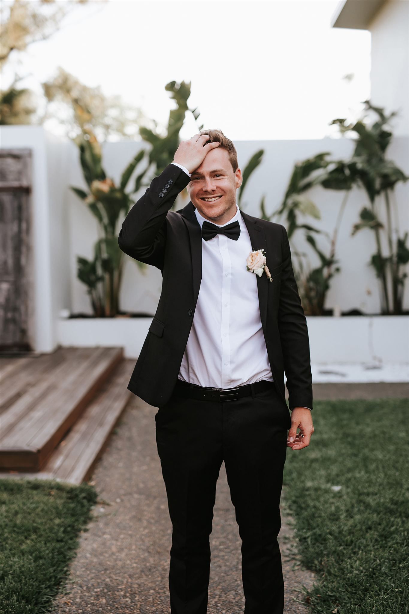 Man in a black suit with bow tie, smiling and touching his hair outside.