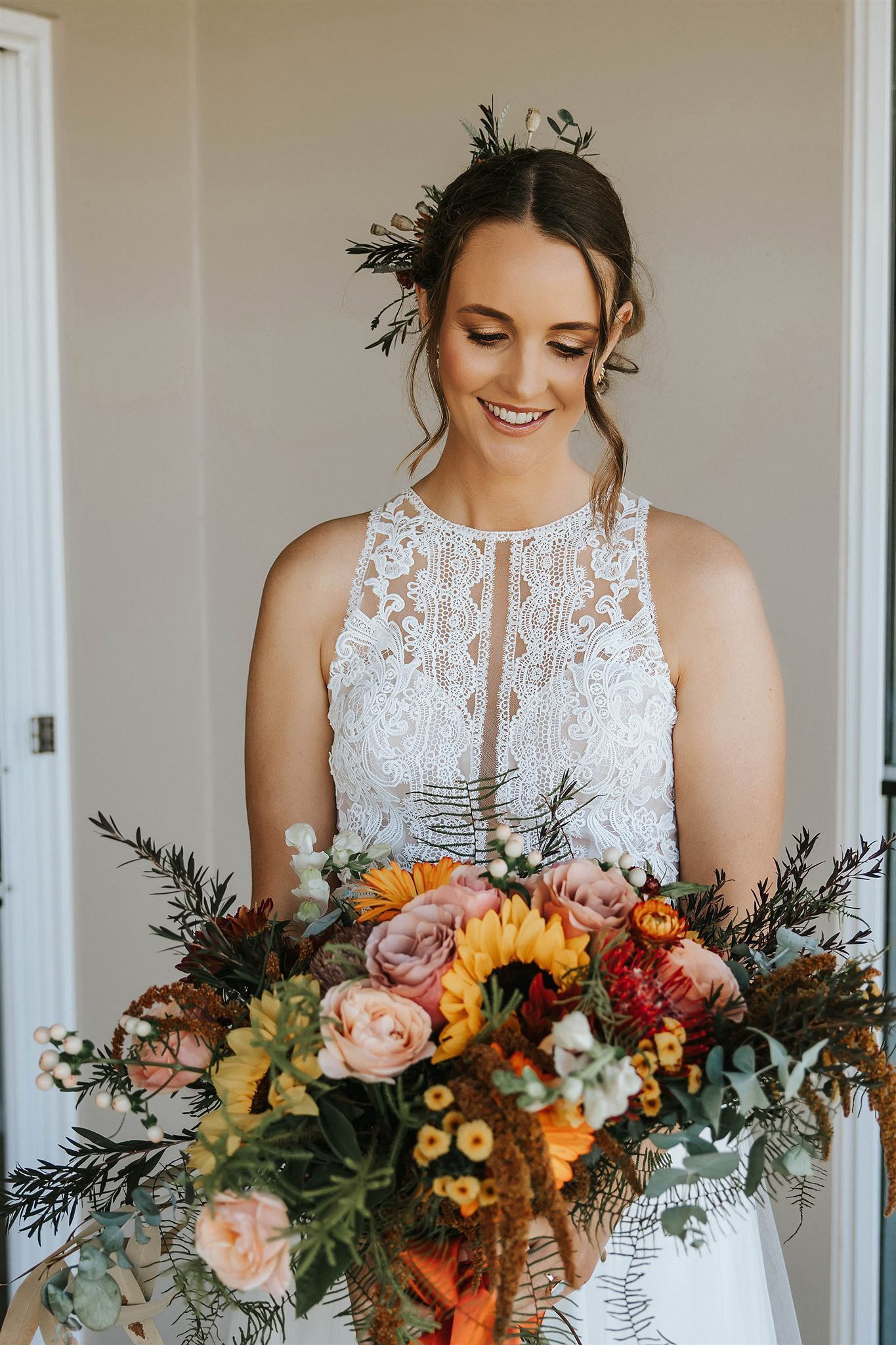 Bride smiles, holding a colorful bouquet. She wears a white lace dress, greenery in her hair, standing indoors.