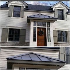 A two-story light gray house with dark shutters and a wooden front door under a metal-roofed entryway.