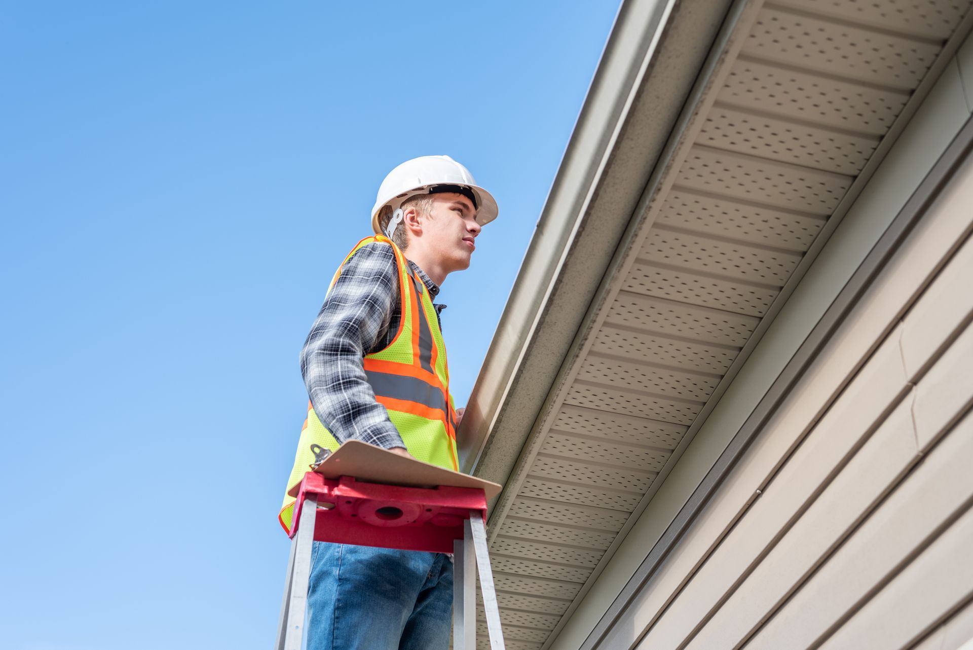 Worker in safety vest and hard hat standing on a ladder near a house roofline