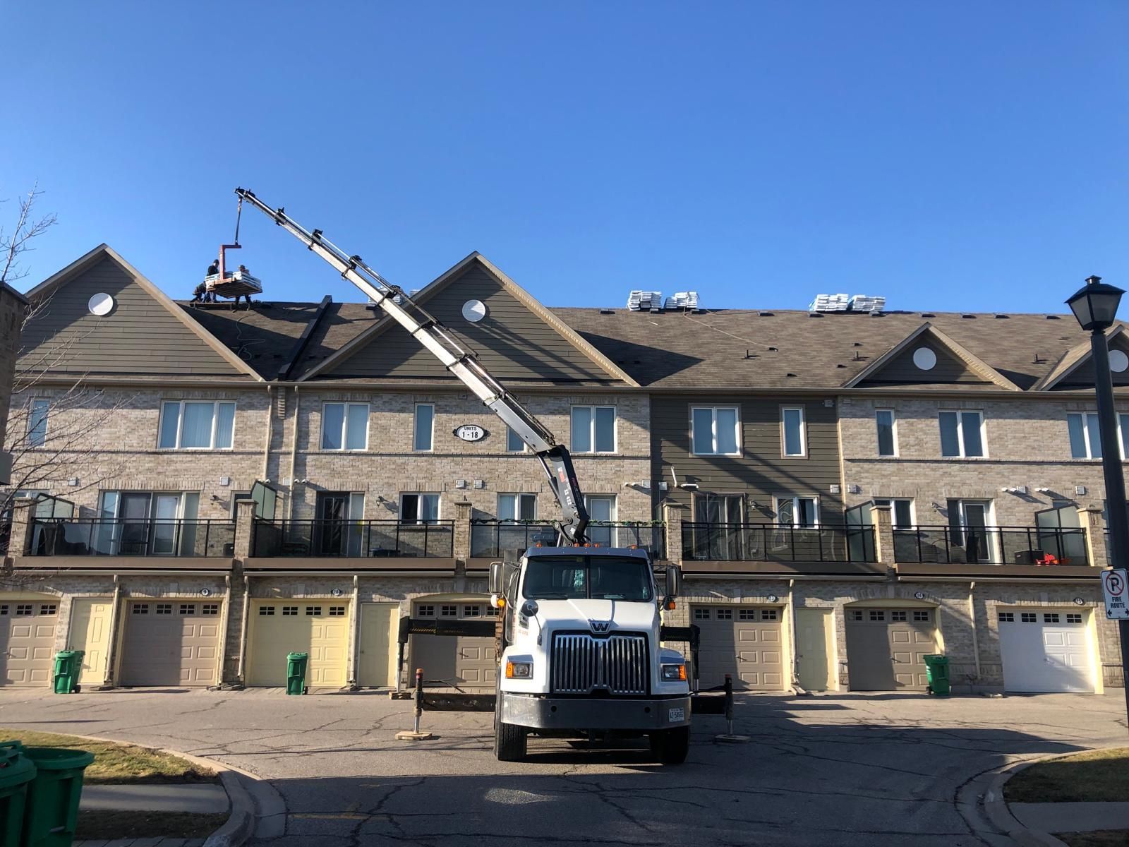 A crane truck parked in front of a multi-unit townhouse complex, lifting materials onto the roof for construction.