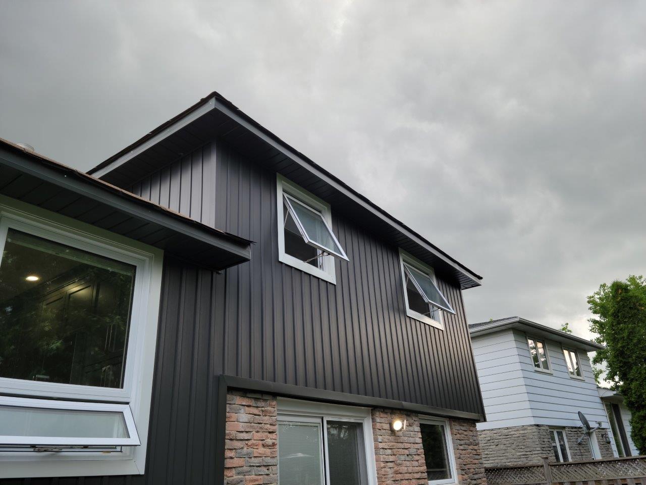 A house with a black siding and white windows is against a cloudy sky.