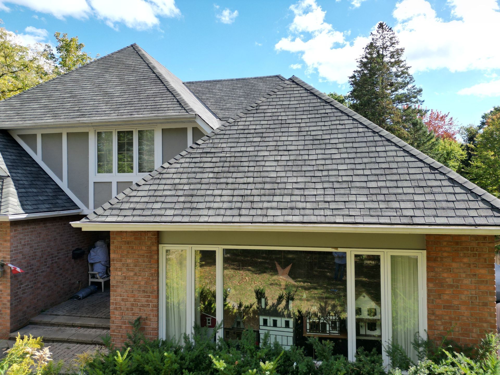 Brick house with gray shingle roof, large front window, and landscaped yard under a blue sky
