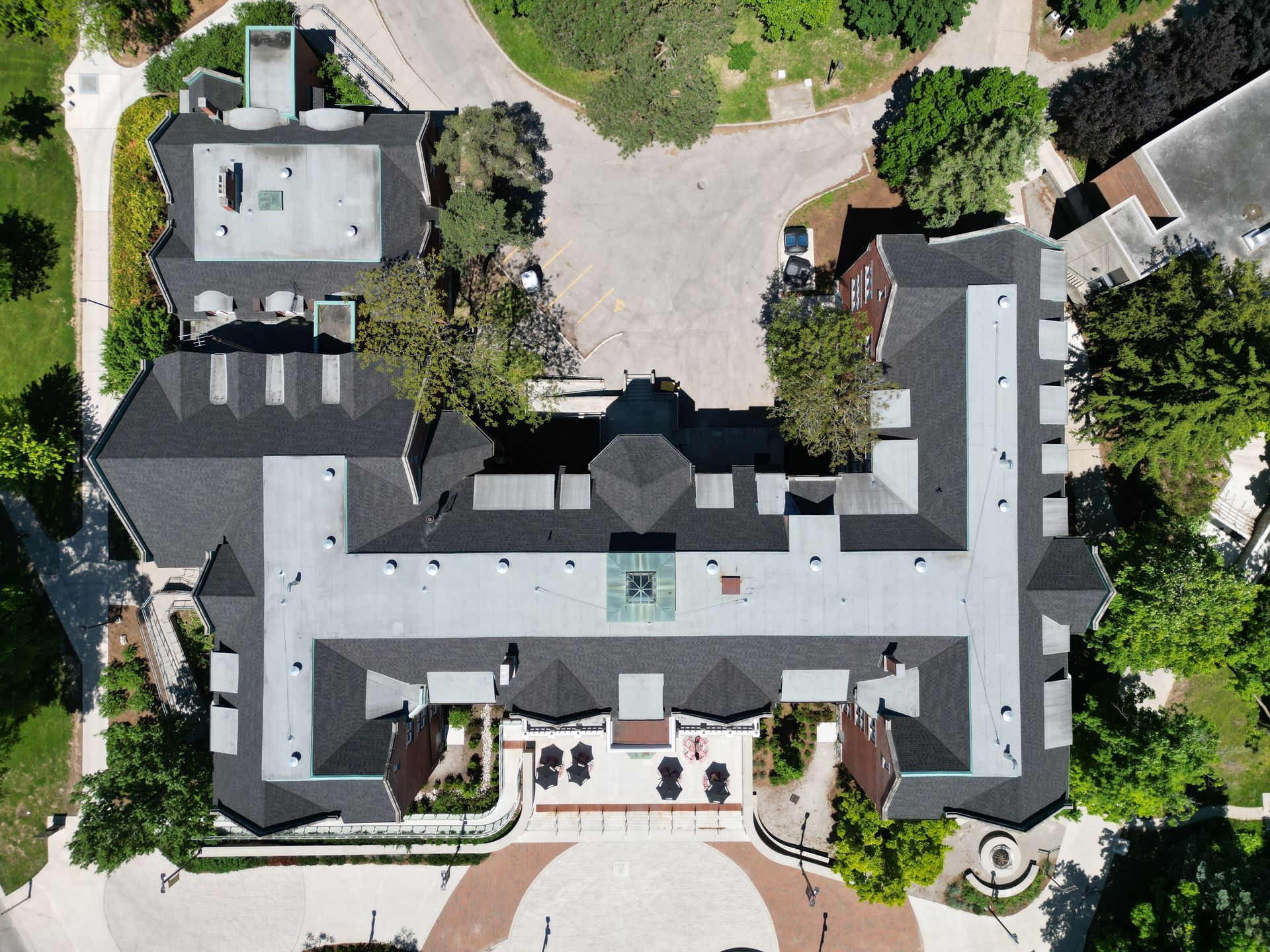 An aerial top-down view of a large, U-shaped brick building with a dark grey roof surrounded by trees and paved walkways.