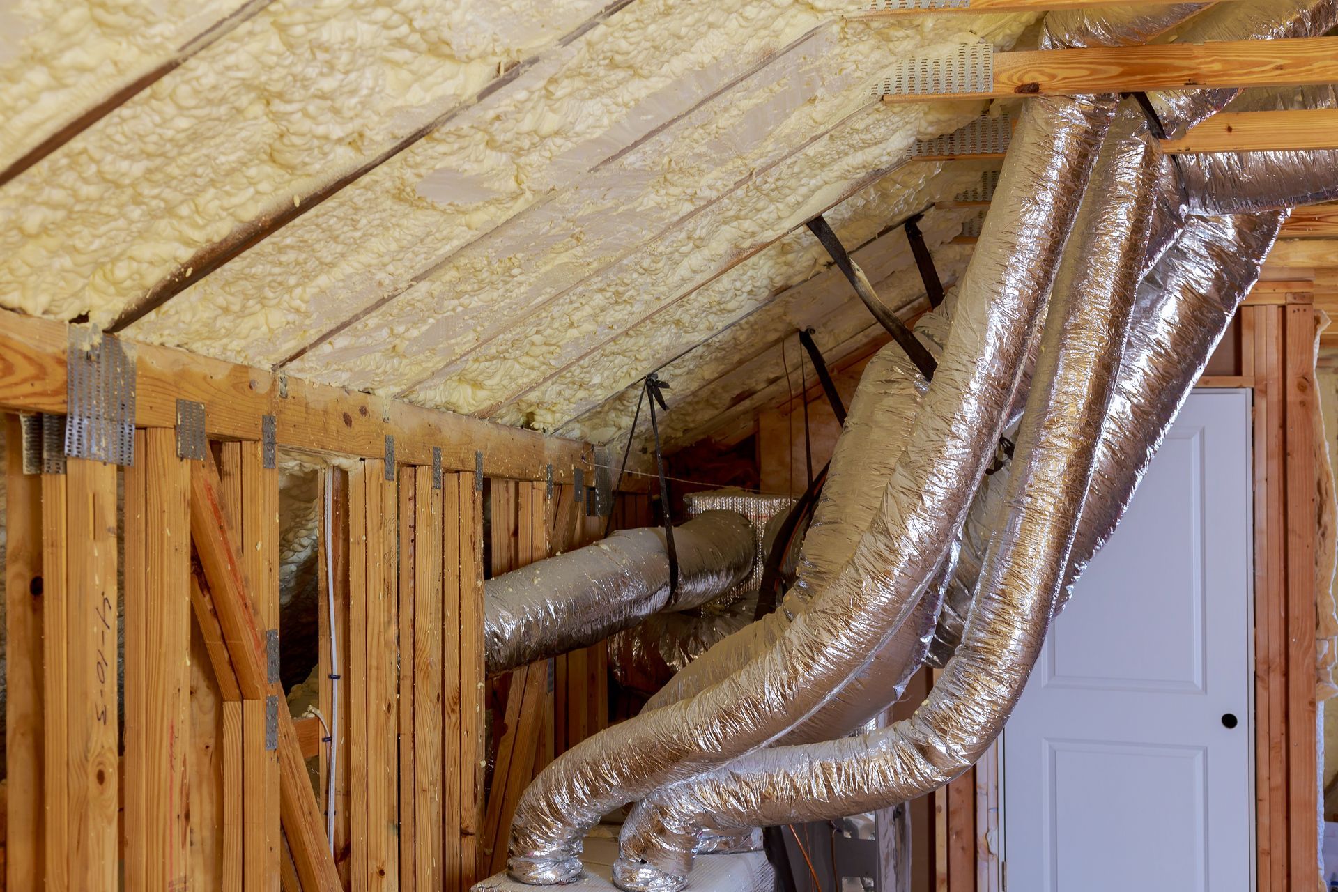 Attic with spray-foam insulation and large silver HVAC ducts above a door